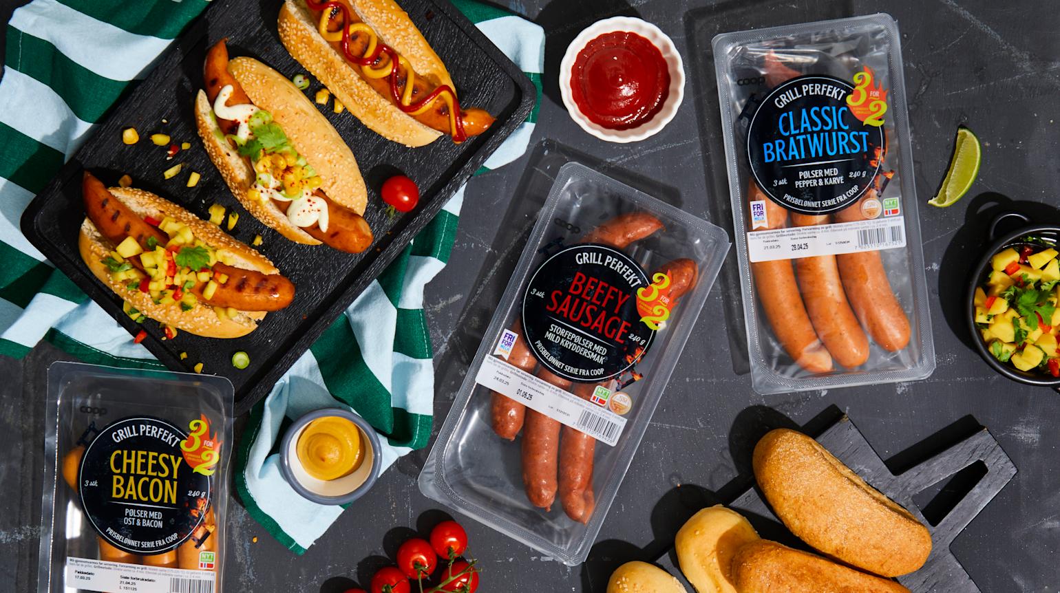 Grill Perfekt pølser i emballasje på mørk steinbakgrunn, samt tilberedte pølser i pølsebrød med forskjellige tilbehør, servert på svart serveringsfat. Mangosalsa, friske cherrytomater og små skåler med tilbehør rundt.