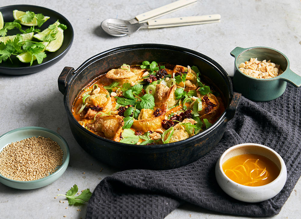 One pan dumplings i red curry saus servet i svart rund ildfast form, med små skåler sesamfrø, appelsinsaus, peanøtter og limebåter ved siden av.