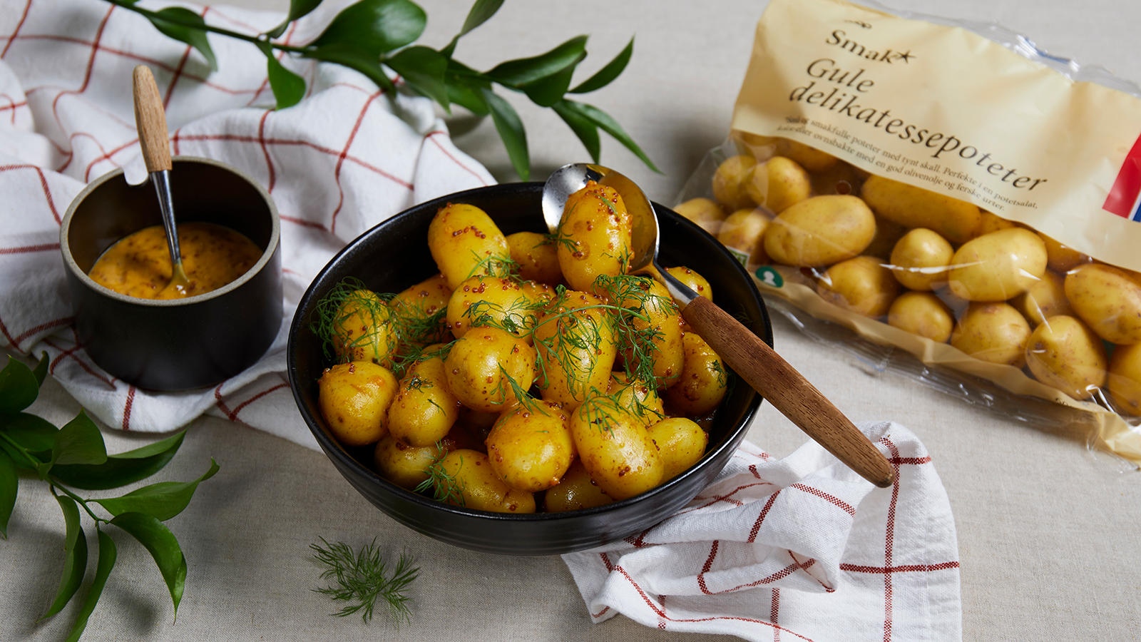 Delikatessepoteter med sennep, honning og dill servert i svart bolle på rød-og hvitrutete kjøkkenhåndkle. Liten skål med sennep ved siden av.