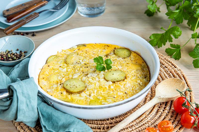 Ekte spansk omelett, med potetskiver og løk, servert i dyp lys stekepanne. Omkranset av røde tomater, grønne urter og lyseblå asjetter på dekket bord.