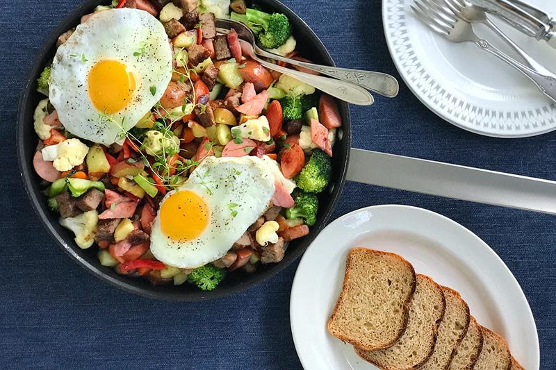 Pytt i panne med forskjellige grønnsaker, pølser, karbonader og spekepølser. Servert i stekepanne med to stekte egg og fersk timian på toppen. Står på middagsbord med et fat brødskiver ved siden av.