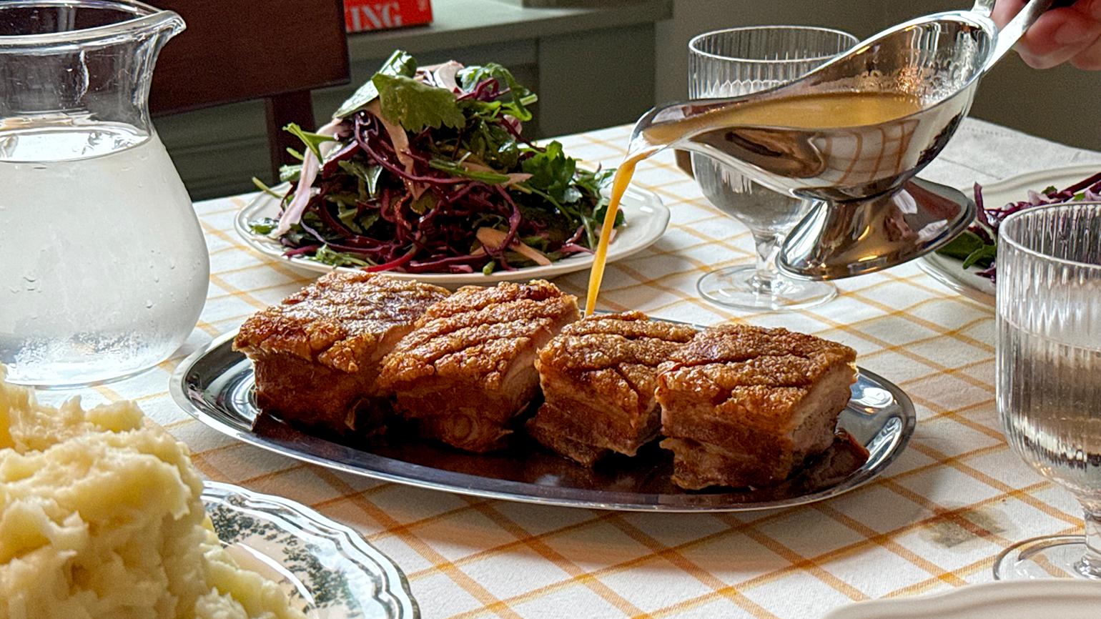 Påskeribbe à la Sultne Gutter på serveringsfat i blankt metall med en asjett rødkål-og fennikelsalat i bakgrunnen. Sjysaus helles over av sausenebb i blankt metall.