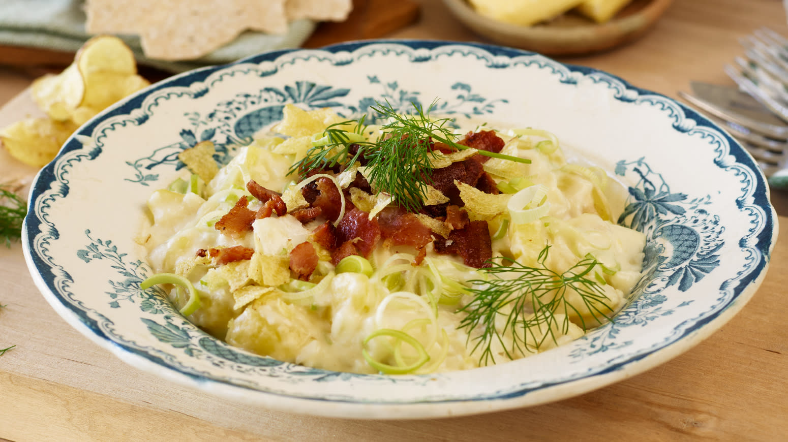 Plukkfisk med bacon og crisp, servert med purre, salt potetgull og frisk dill i blåmønstret hvit skål på dekket middagsbord.