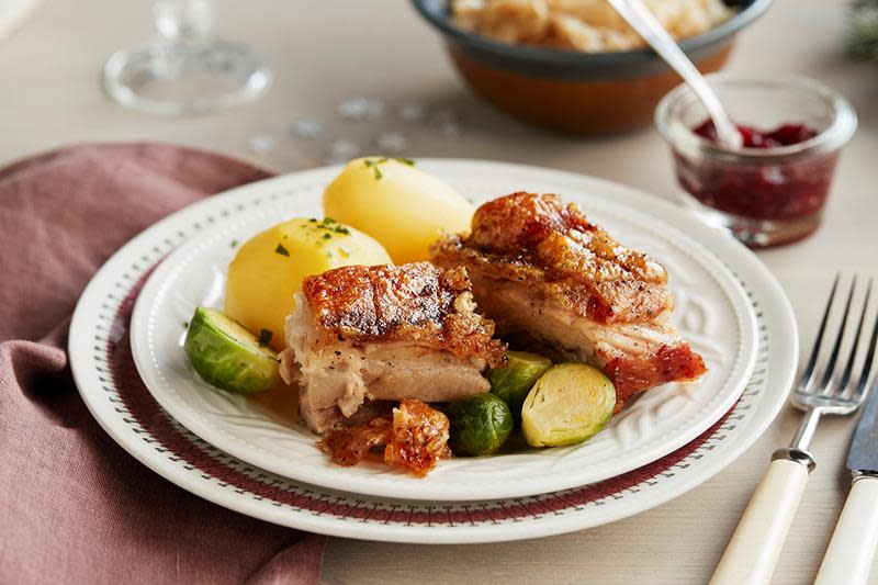 Gyllen ribbe med sprø svor, servert i biter på hvit tallerken med kokte poteter og rosenkål. Tallerkenen står på dekket middagsbord med skåler med tyttebær og surkål i bakgrunnen.