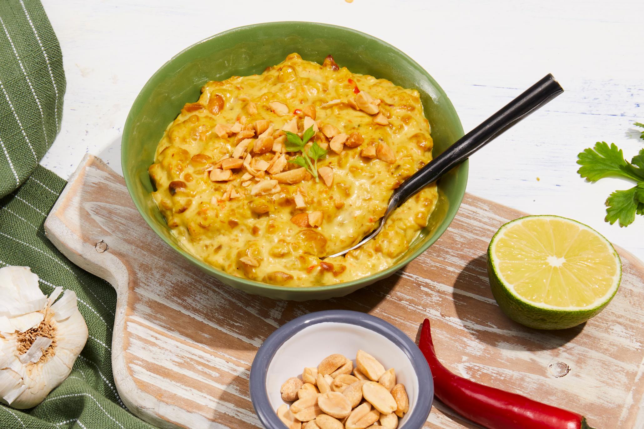 Sataysaus til kylling servert i grønn skål på lys trefjøl. Halv lime, hel rød chili, hvitløk og en liten skål peanøtter rundt.
