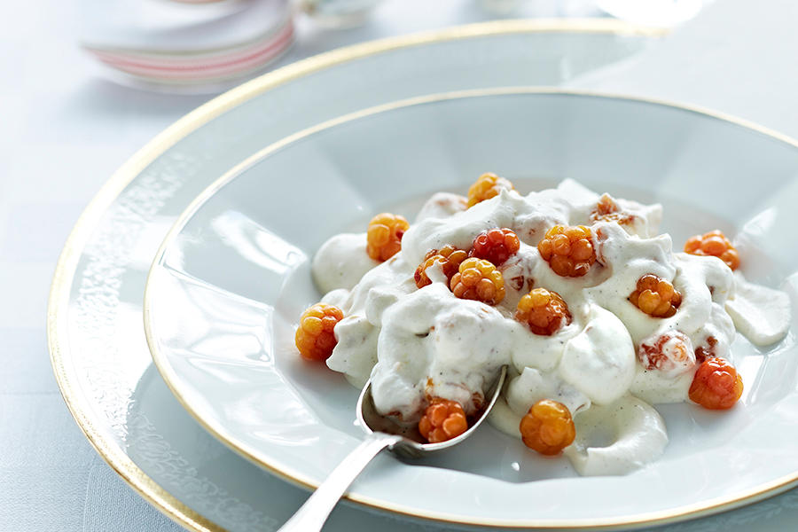Hjemmelaget multekrem med vanilje, servert i gullkantet dessertskål på bord med hvit duk. Multekremen er pyntet med friske multer, og har en sølvskje stukket inn.