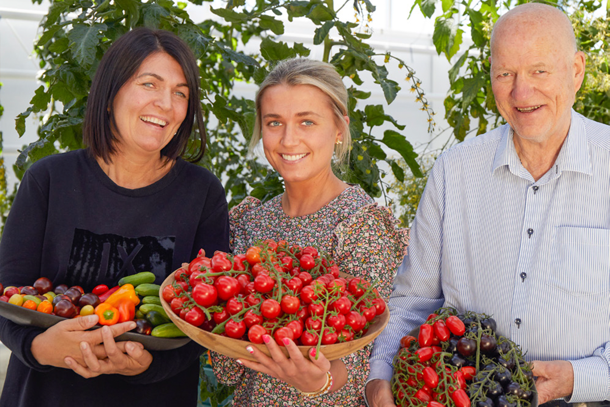 Tre personer står smilende med tomater og andre grønnsaker i hendene