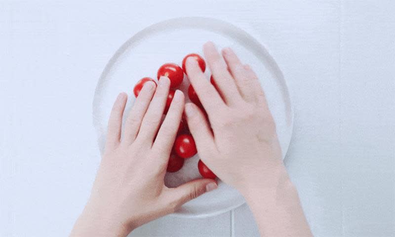 Hvit rund tallerken med flere røde små tomater på hvit bakgrunn. To hender holder fast tomatene.