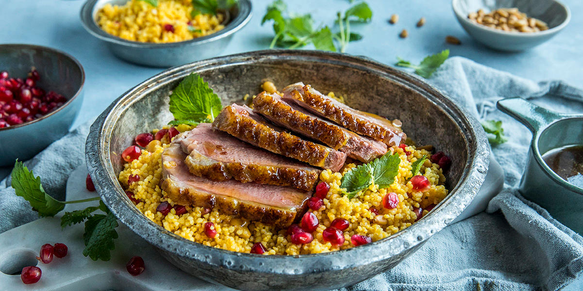 Marokkansk andebryst med couscous og appelsinsaus servert i rustikk metallskål med granateplefrø og ferske urter. Skålen står på lyseblått bord med små skåler couscous, granateplefrø, pinjekjerner og saus.