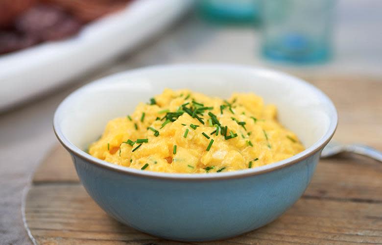 Eggerøre drysset med finhakket gressløk i blå skål, servert på trefjøl. Frokostbord i bakgrunnen.