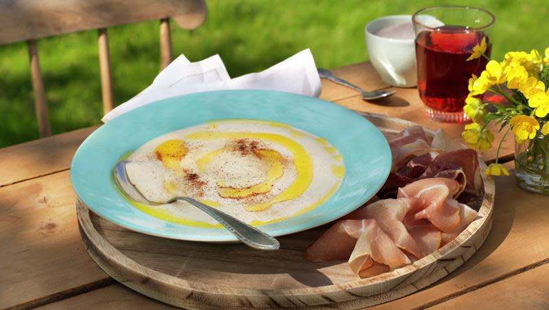 Hjemmelaget rømmegrøt drysset med sukker og kanel, og servert i turkis dyp tallerken med smeltet smør. Tallerkenen står på trefat med spekemat, på et hyggelig pyntet sommerbord i hagen. Glass med rød saft ved siden av.