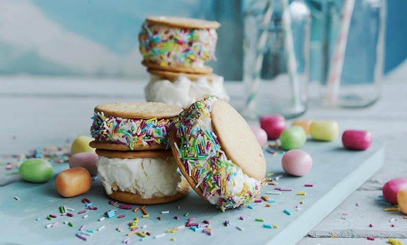 Iskrem-sandwich med kun 3 ingredienser, pyntet med tutti frutti kakestrø, ligger på lys blå trefjøl med godteri rundt.