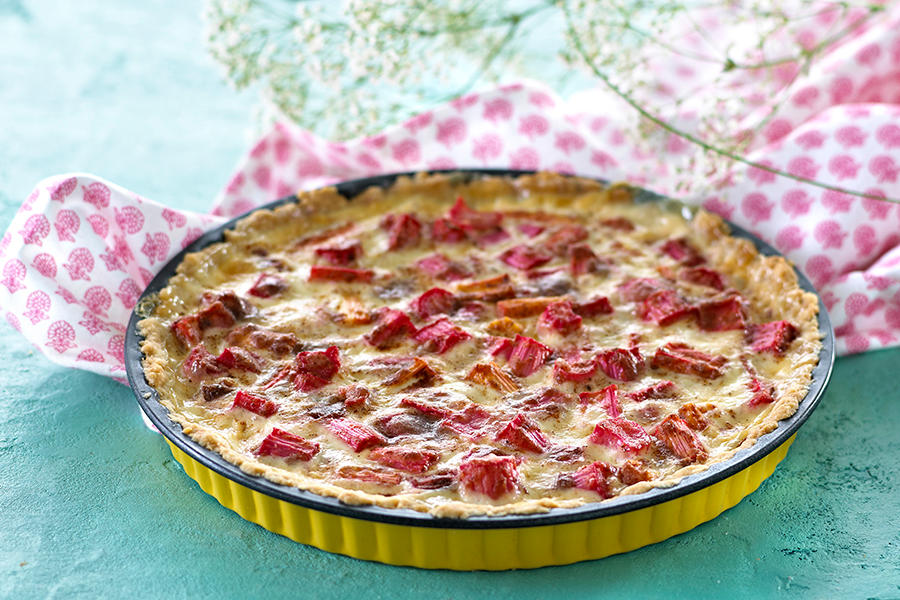 Rabarbrapai i gul paiform, servert på turkist underlag med rosamønstret kjøkkenhåndkle ved siden av. Stilk med hvite blomster i bakgrunnen.