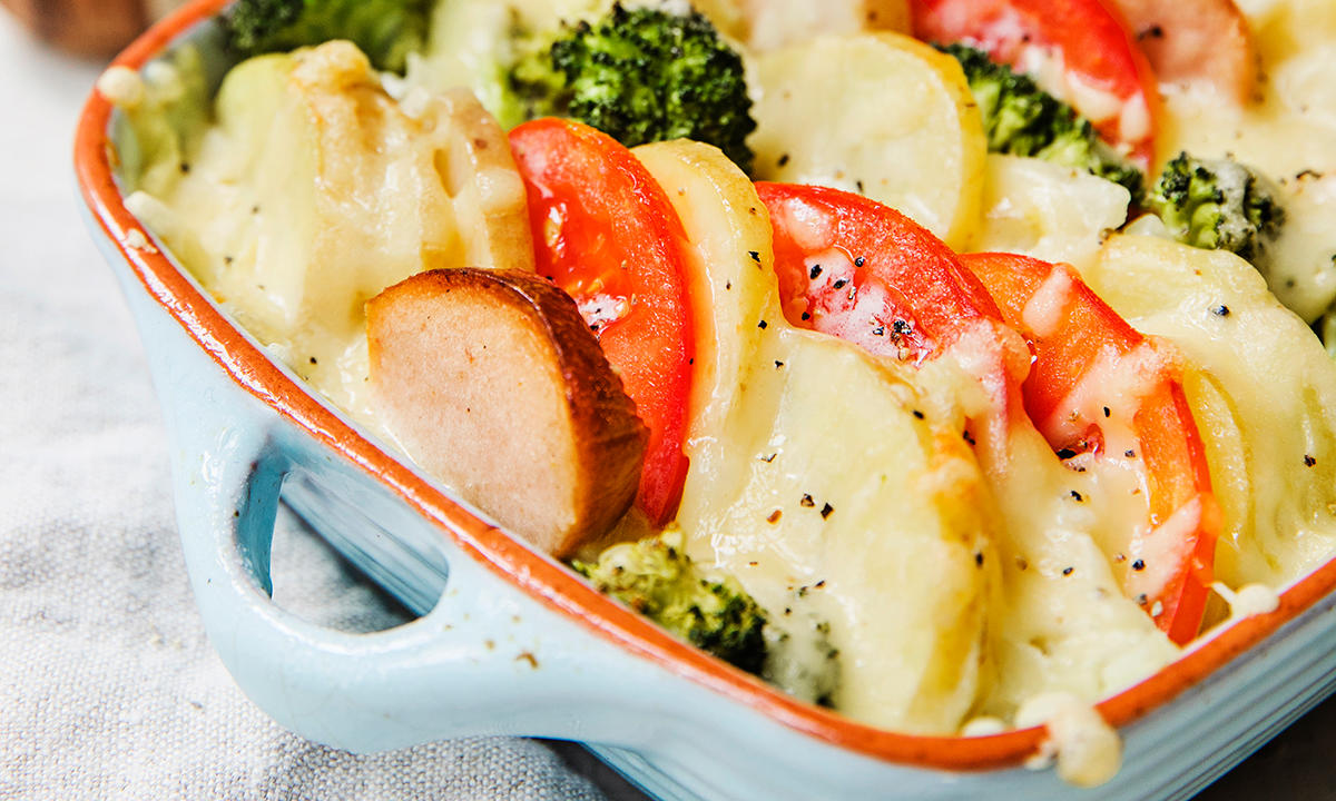 Pølsegrateng med brokkoli og potet, og tomat i skiver, servert i lyseblå ildfast form.