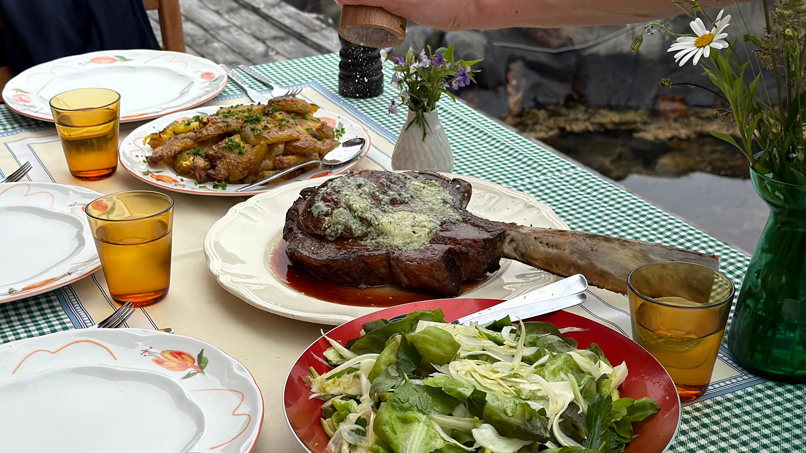 Grillet tomahawk med blåmuggostsmør servert på fat på hagebord med grønnrutet duk og gule glass. Fat med stekte poteter og bolle med grønn salat ved siden av.