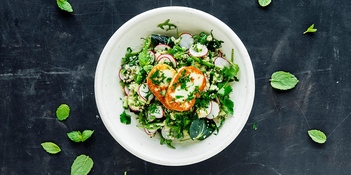 Cousoussalat med halloumi og sitronvinaigrette, redikk og ferske urter servert i hvit bolle på svart marmorbakgrunn.