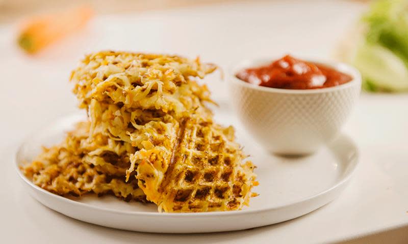 Hash browns, eller røstipoteter, med blomkål og gulrot stekt i vaffeljern. Stablet på hvit tallerken, med ketchup i hvit skål ved siden av.