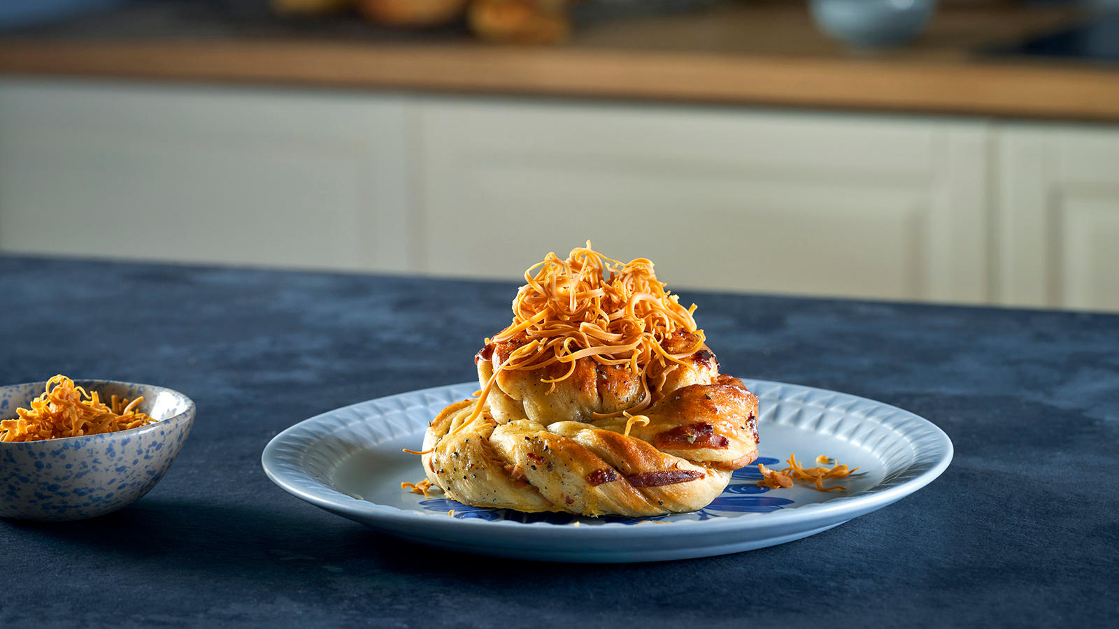 Kanelboller med brunost på toppen servert på lyseblå asjett med en liten skål revet brunost på siden.