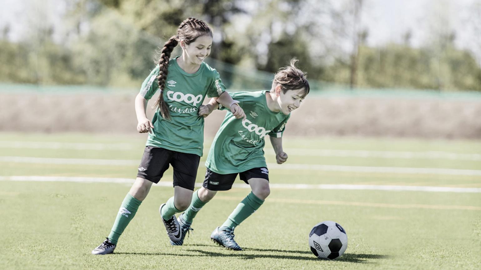 En gutt og en jente i grønne fotballtrøyer med Coop- logo, og svarte fotballshorts spiller fotball på grønt underlag.