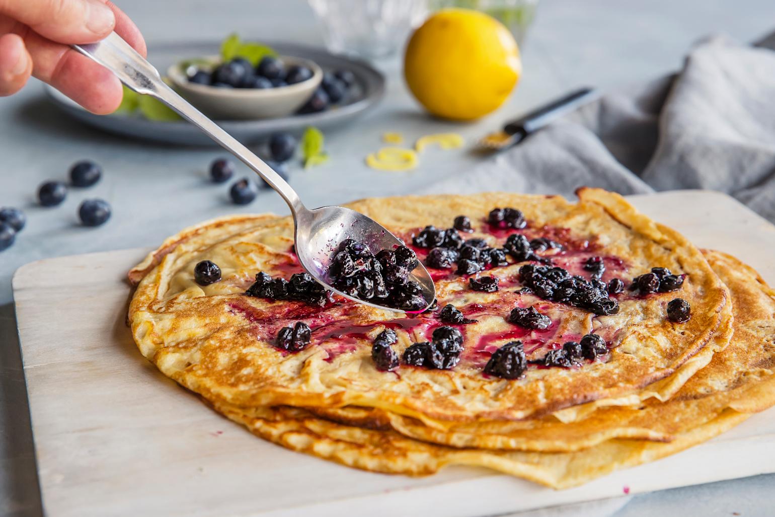 Pannekaker med hjemmelagde rørte blåbær på toppen servert på lys trefjøl. I bakgrunnen står det en skål friske blåbær, og en gul sitron. En hånd holder en skje med rørte blåbær over pannekakene.