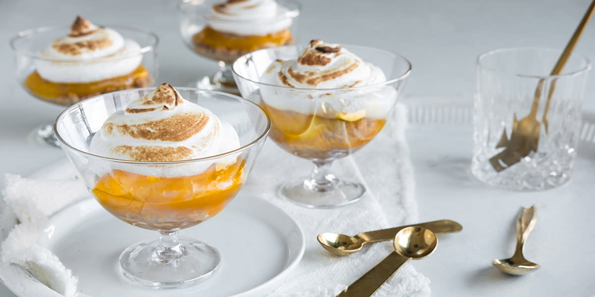 Lemon meringue pie på glass servert lagvis i dessertskåler med stett, på hvitt serveringsbrett med hvit linserviett og gullfargede teskjeer.