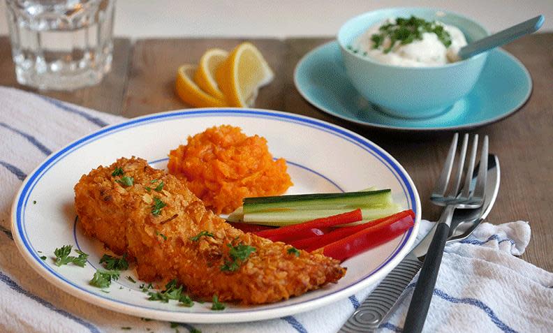 Cornflakespanert laks med søtpotetmos, agurkstenger og paprikastenger på hvit tallerken. Hvit urtedip i blå skål i bakgrunnen.