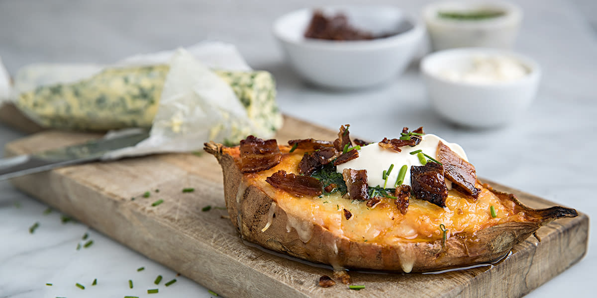 En halv bakt søtpotet med bacon og urtesmør servert på trefjøl med en rull hjemmelaget urtesmør ved siden av. I bakgrunnen er tre små hvite skåler med sprøstekt bacon, rømme og gressløk.