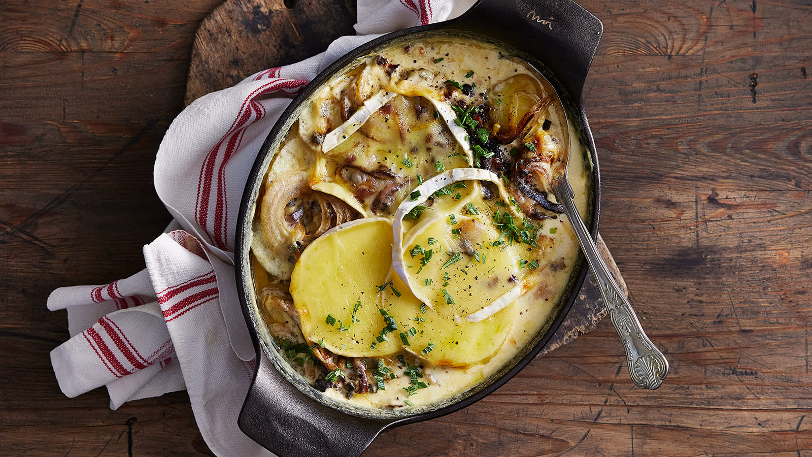 Tartiflette - fransk potetgrateng med brie servert i mørk ildfast støpejernsform på rustikk trebord.