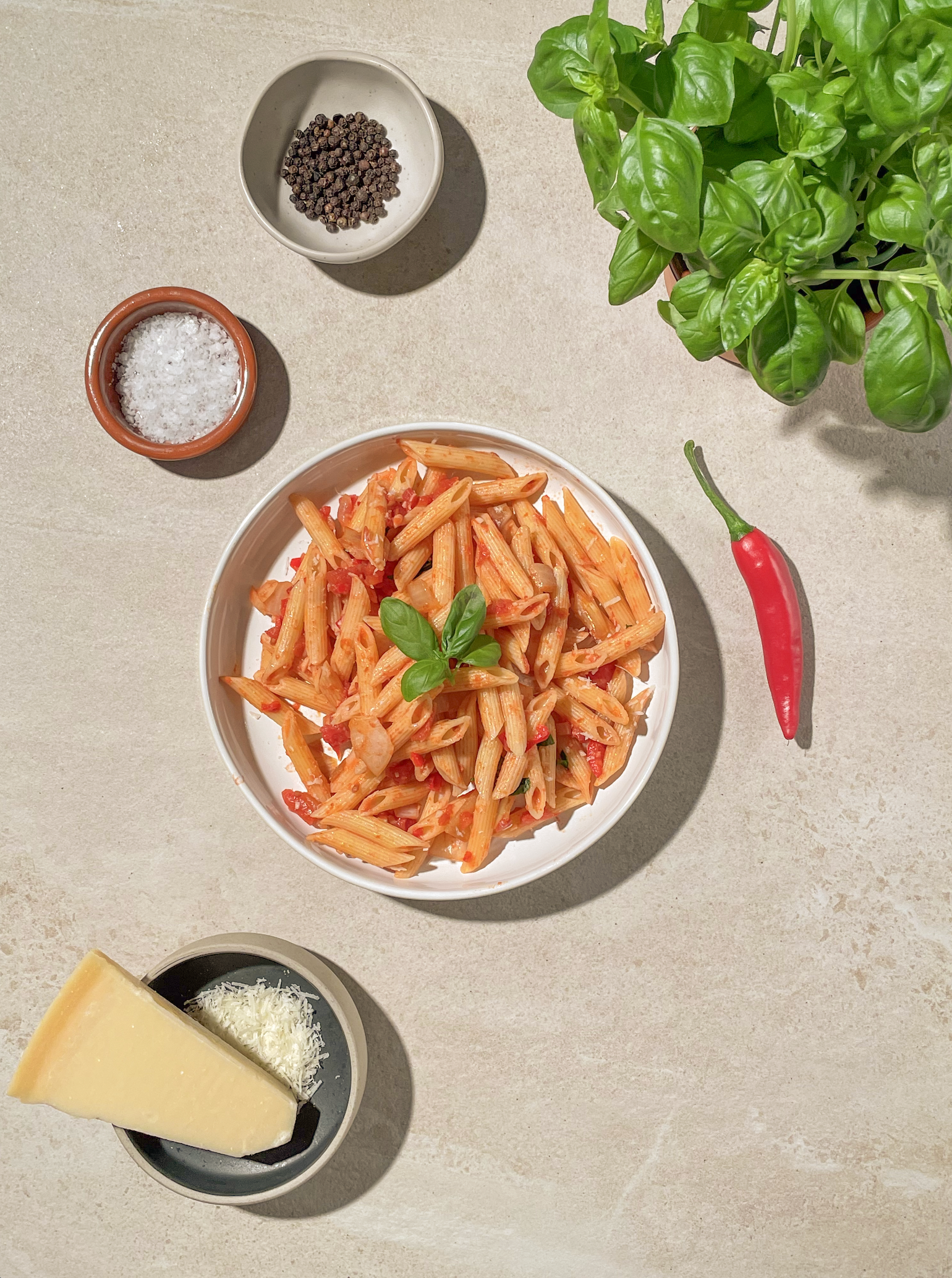 Pasta arrabiata i hvit skål, på lys bakgrunn. Fersk basilikum, chili, parmesan, salt og pepper rundt.