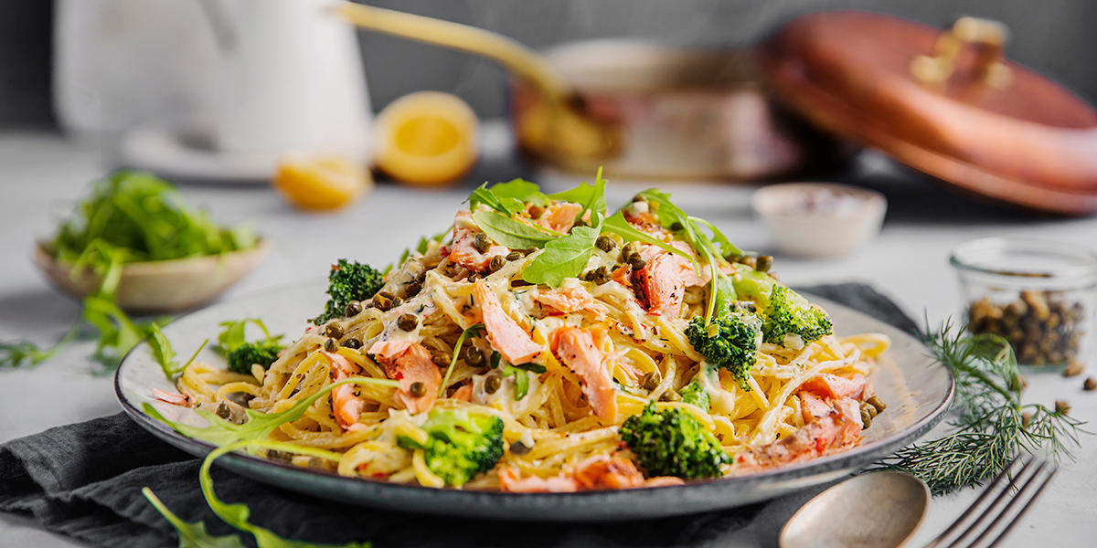 Kremet pasta med varmrøkt laks, sitronpepper og kapers, servert på lys keramikktallerken med brokkoli og ruccolasalat. Messingpanne, skål med kapers, skål med ruccola og frisk sitron i bakgrunnen.