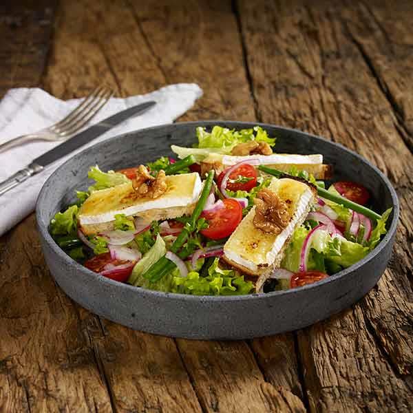 Salat med gratinert brie, valnøtter, tomat, og rødløk servert i mørk grå skål på rustikk trebord.