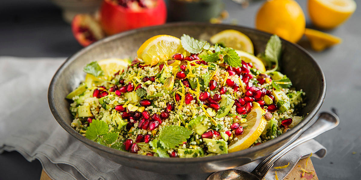 Couscoussalat med granateplekjerner, pistasjnøtter, revet sitronskall, avokado og urter servert i mørk bolle.