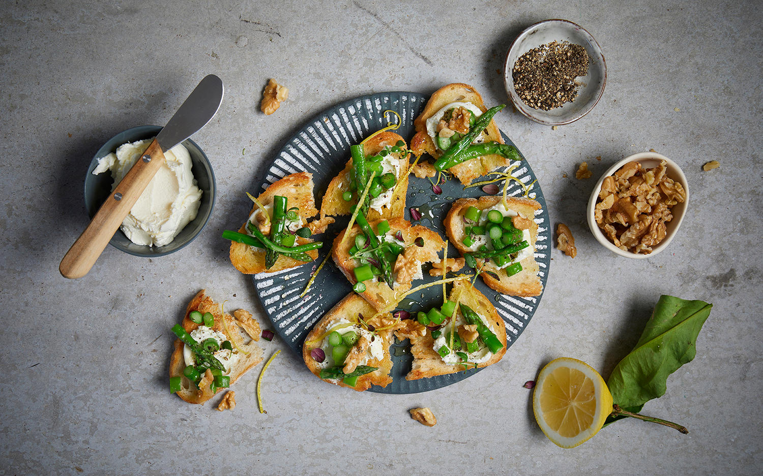 Bruschetta med kremet geitost, asparges og valnøtter servert på mørk tallerken med flytende honning, frisk timian og revet sitronskall.