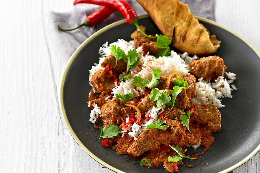 Indisk lam korma med ris og naanbrød, servert på mørkegrå tallerken med et dryss koriander over. Hele røde chili på siden.