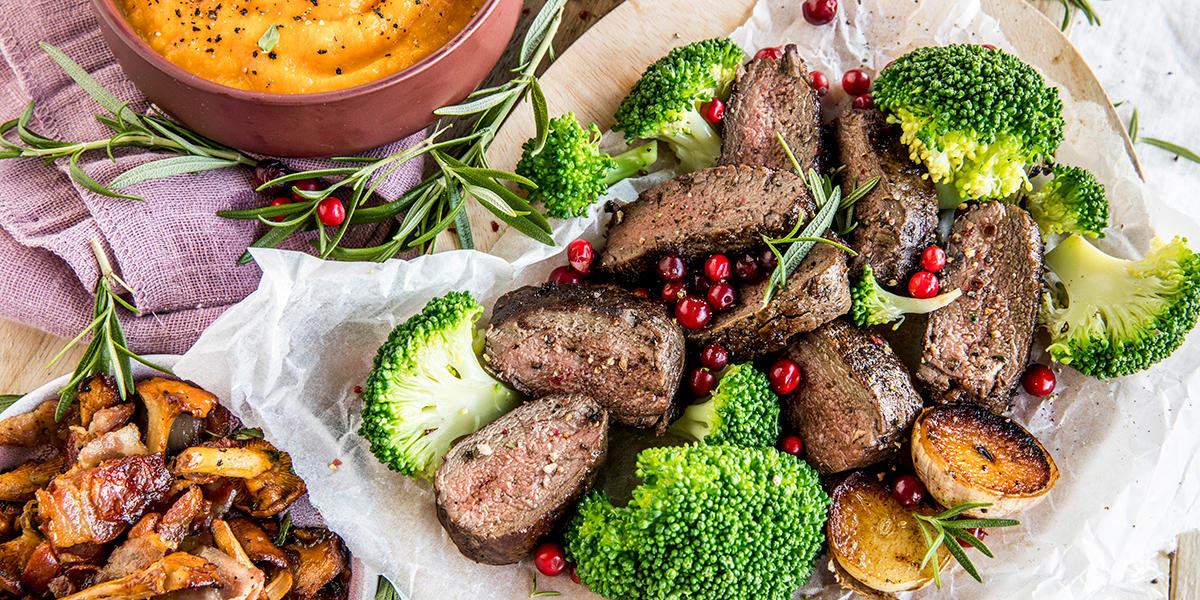 Reinsdyrfilet med smørstekte kantareller, bacon og søtpotetpuré servert på hvitt bakepapir med brokkoli.