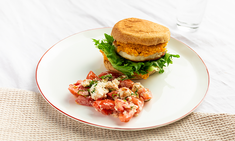 Fiskeburger med hummus og tomater servert på en hvit tallerken med rød kan, ligger på en hvit duk og et beige håndkle