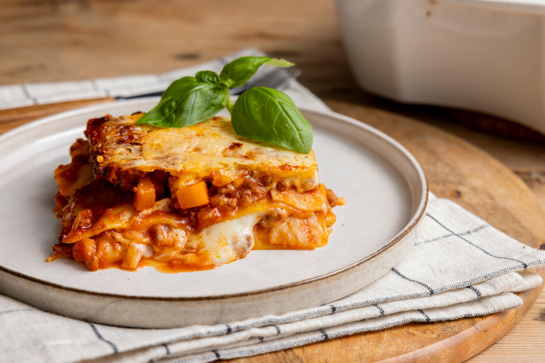 Enkel lasagne med høstens rotgrønnsaker laget av influenser Warunee, servert i stykke på hvit tallerken med frisk basilikum på toppen. Tallerkenen står på rund trefjøl med stripete kjøkkenhåndkle under.