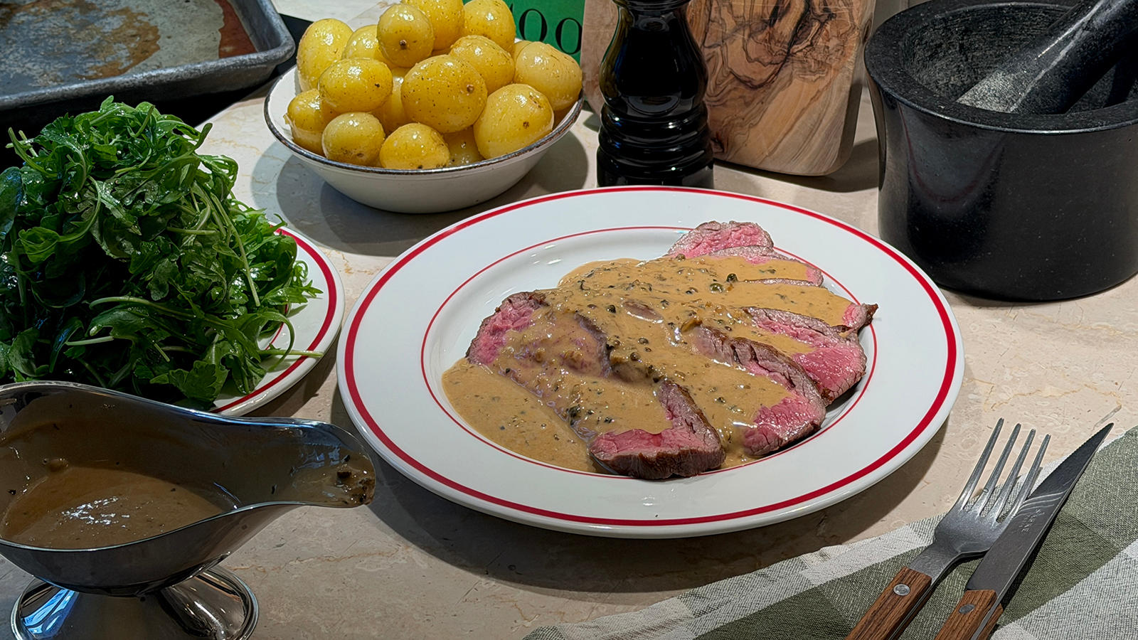 Flankestek av limousin med trippel peppersaus servert på hvit tallerken med rød kant, med sausenebb i forkant, og to skåler tilbehør i bakgrunnen.