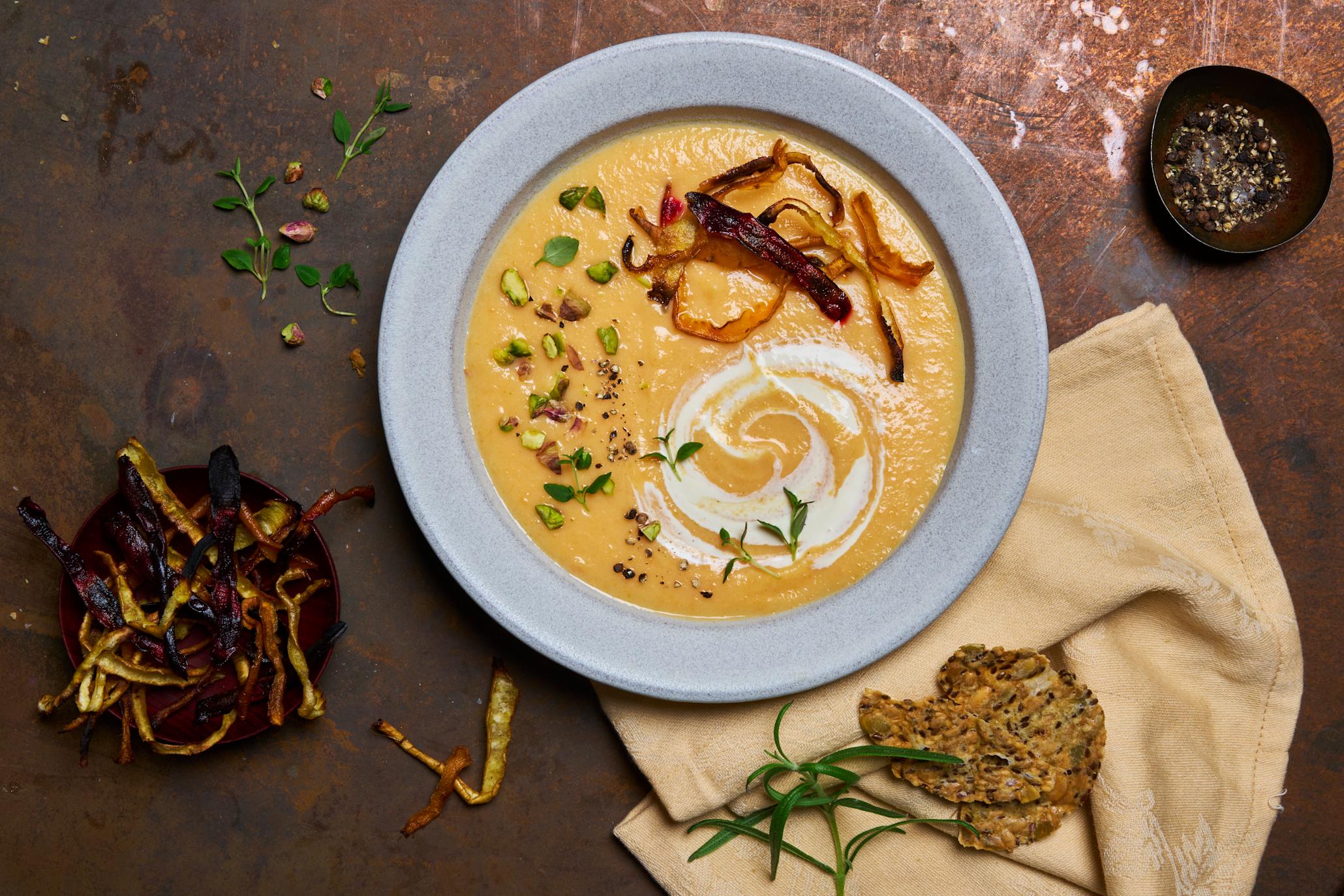 Grønnsakssuppe med rotgrønnsaker og urteknekkebrød servert i en lyseblå skål, med linserviett og knekkebrød ved siden av.