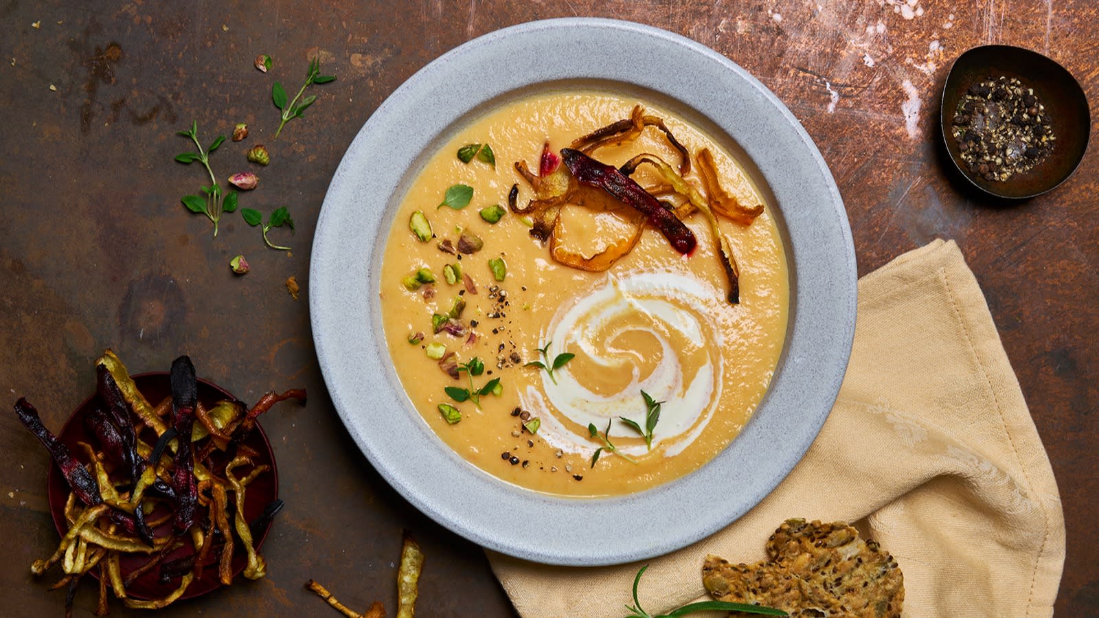 Grønnsakssuppe med rotgrønnsaker og urteknekkebrød servert i en lysegrå skål, med linserviett og knekkebrød ved siden av.