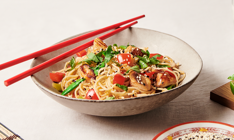 Rød paprika, sukkererter og kylling over eggnudler dandert med hvite og svarte sesamfrø i en grå skål med et par røde spisepinner.