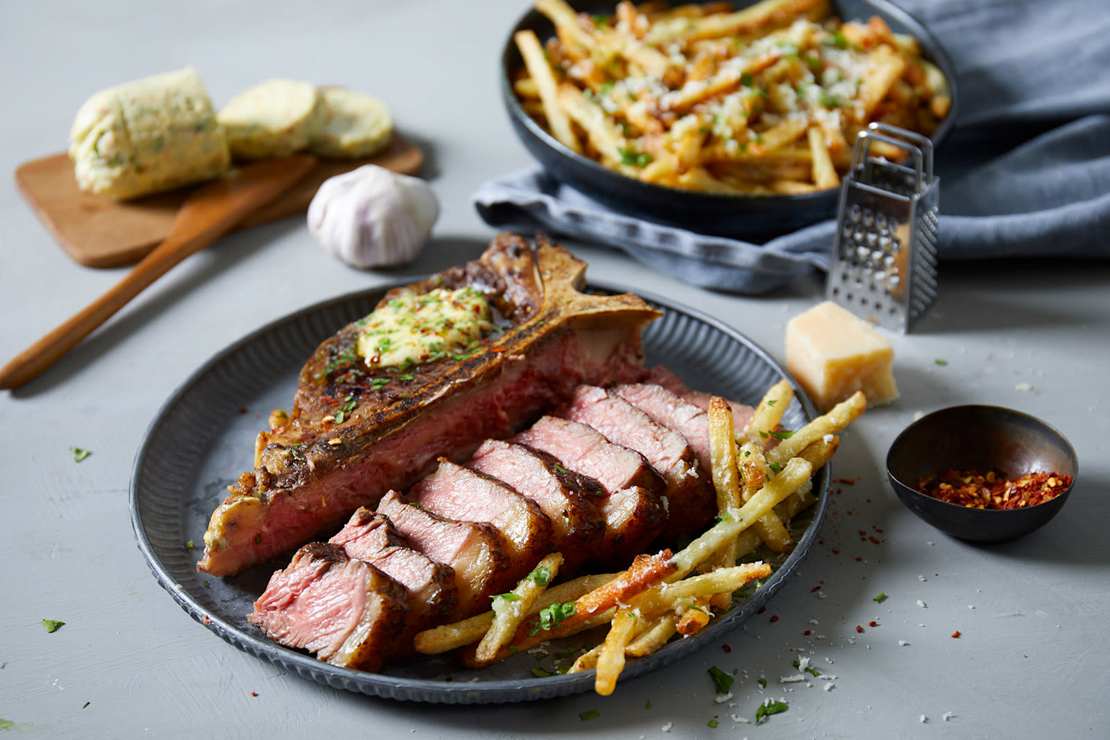 T-Bone steak med cowboy butter og parmesan fries servert på mørkegrå tallerken med en liten skål chiliflak på siden. Liten fjøl med kryddersmør, og skål med fries i bakgrunnen.