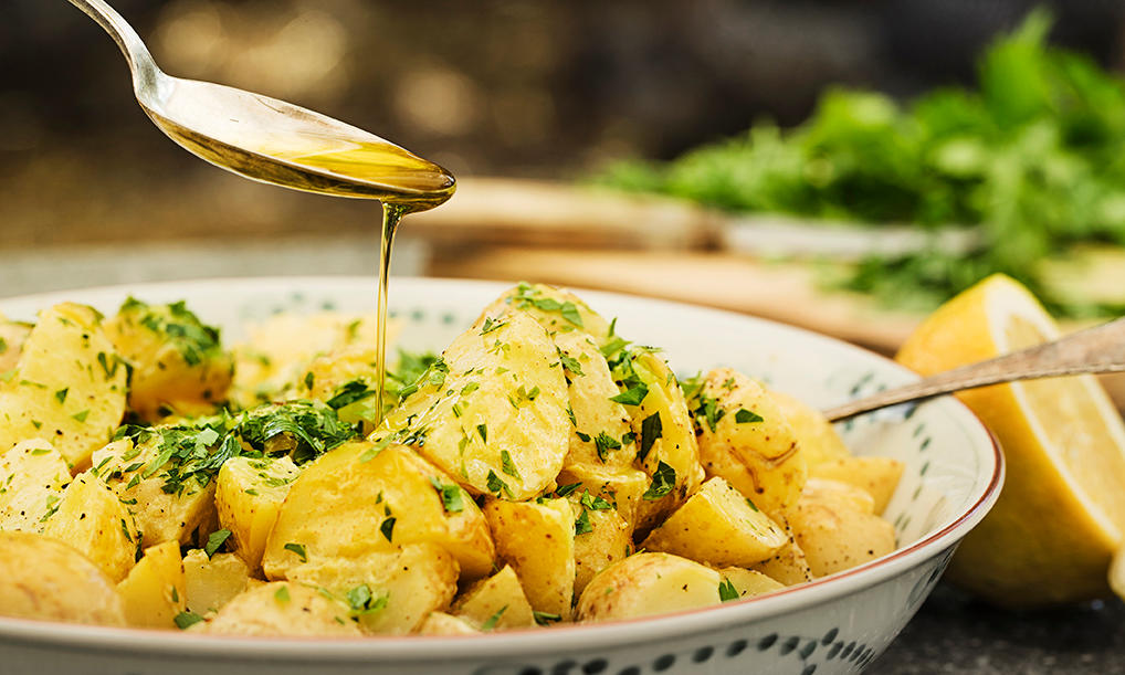 Potetsalat med fransk sennepsdressing servert i lys skål med fersk sitron og urter på siden. Sennepsdressing helles over med spiseskje.
