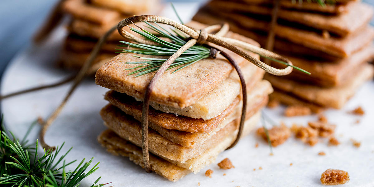 Knasende sprø kolakaker; firkantede tynne småkaker, stablet i små tårn, bundet sammen med hyssing og pyntet med litt granbar. Stablene er servert på rund hvit steinfjøl.