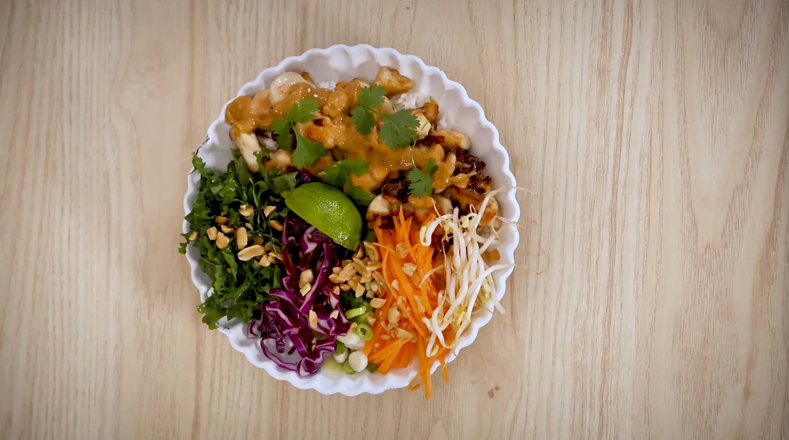 Kyllingbowl med sataysaus, finkuttede grønnsaker og hakkede peanøtter servert i hvit bolle på lyst trebord, med en båt lime på toppen.