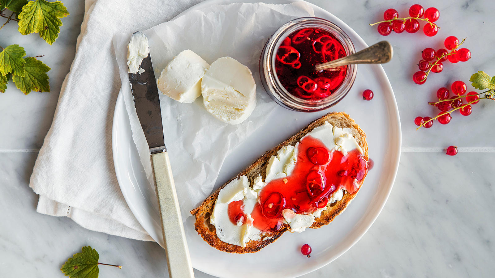Hjemmelaget ripsgelé med chili servert på brødskive med chevre på hvit tallerken. Syltetøyglass med ripsgele ved siden av, og biter av chevre på tallerkenen.