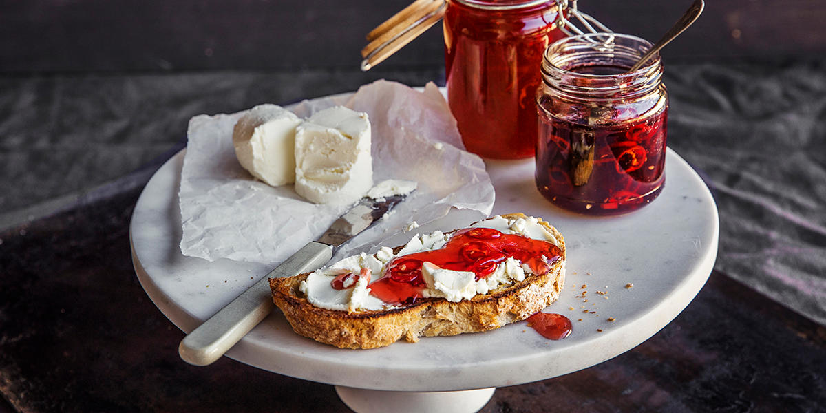 Hvitt stettefat med et glass med ripsgele, et glass med ripsgele med chili, en ristet brødskive med chevre og ripsgele og biter av chevre på matpapir.
