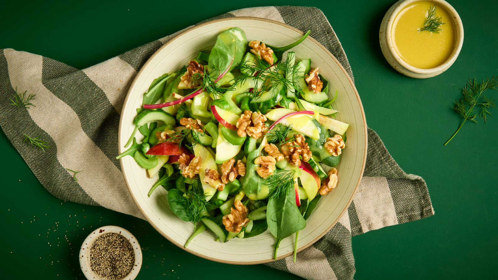 Høstsalat med agurk, eple, selleri og valnøtter servert i beige skål på grønt- og naturstripet kjøkkenhåndkle. Liten mugge med sennepsvinaigrette på siden.