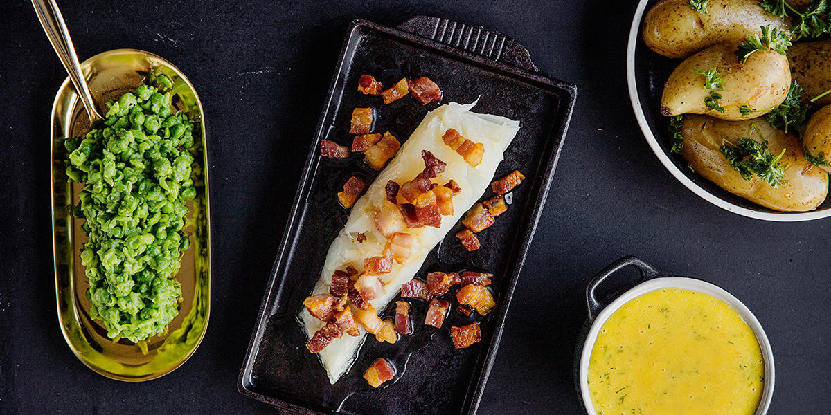 Lutefisk i ovn med sennepssaus og ertestuing servert på svart støpejernsfat med sprøstekt bacon på toppen. Fatet er omkranset av flere små skåler med henhodsvis sennepssaus, kokte persillepoteter og hjemmelaget ertestuing.