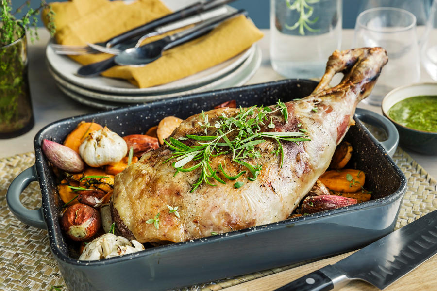 Helstekt lammelår i ildfast form, toppet med friske urter. Ovnsbakte grønnsaker og hele hvitløk i bunnen av formen. Myntepesto i liten skål på siden.